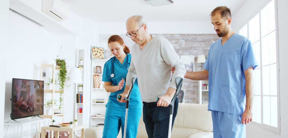 Team of nurses or social workers helping an old disabled man to walk with his crutches out of the nursing home room Team of nurses or social workers helping an old disabled man to walk with his crutches out of the nursing home room.