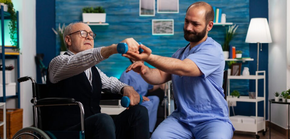 Practitioner man doctor helping retiree senior male in wheelchair to do physiotherapy strength exercise Practitioner man doctor helping retiree senior male in wheelchair to do physiotherapy strength exercise using dumbbell. Social nursing services at home. Healthcare recovery. Support worker helper