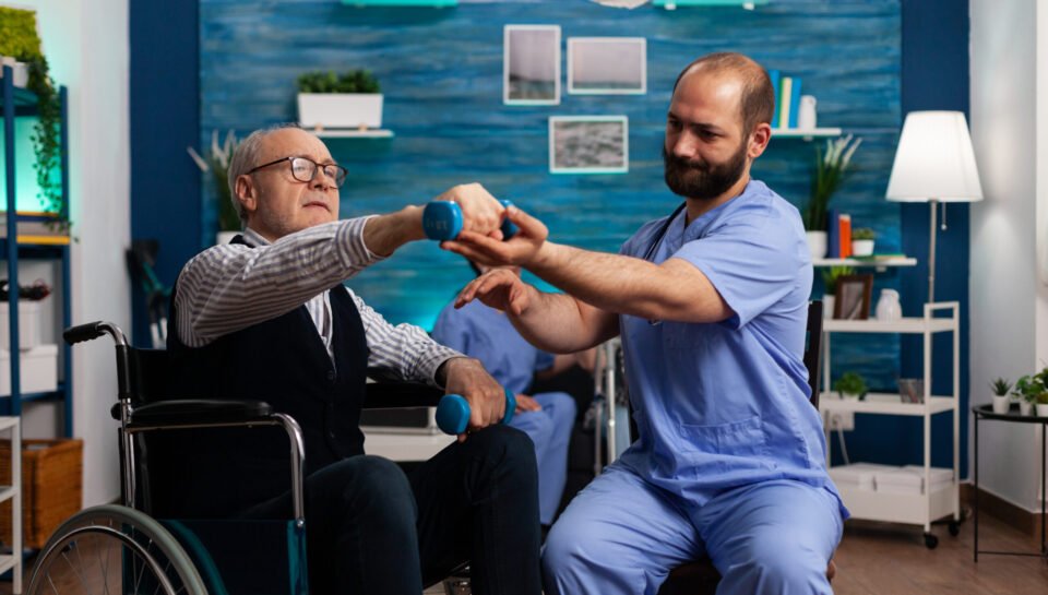 Practitioner man doctor helping retiree senior male in wheelchair to do physiotherapy strength exercise using dumbbell. Social nursing services at home. Healthcare recovery. Support worker helper